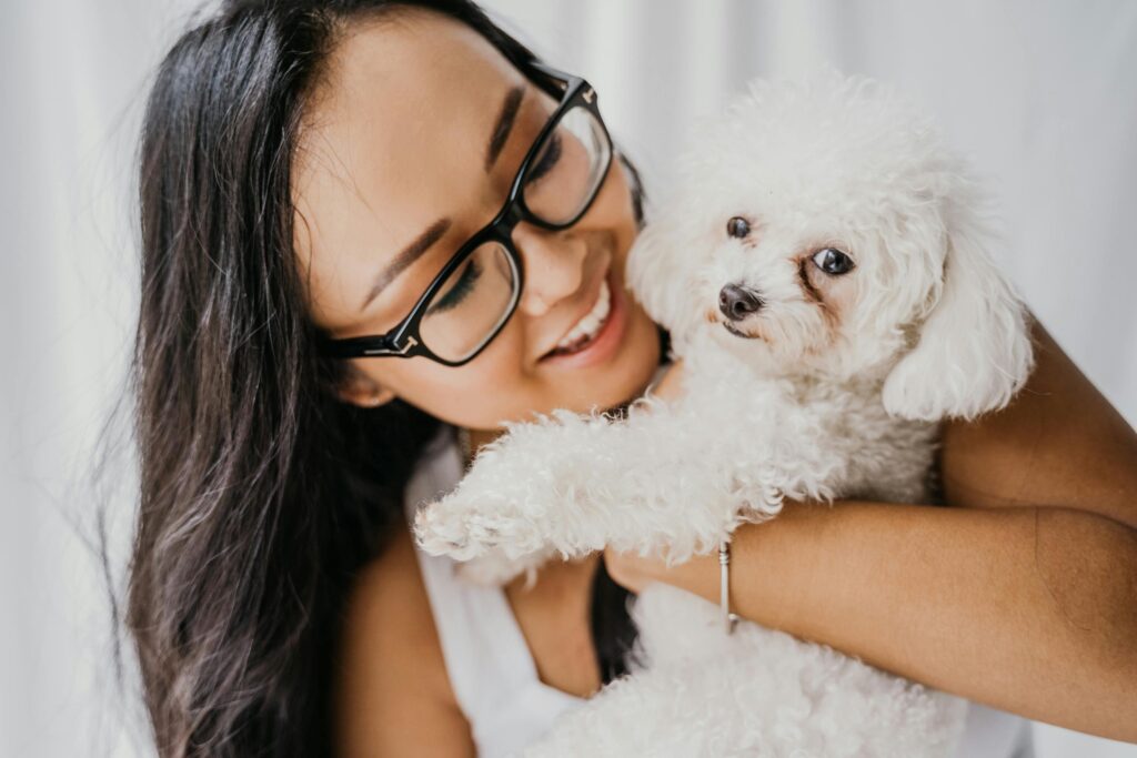 Close-up of a smiling Asian woman with eyeglasses hugging a white poodle indoors. Warm and joyful moment captured.