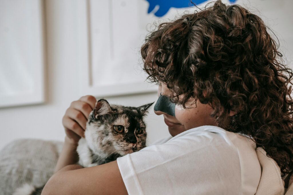 A woman smiling indoors while cuddling her furry pet cat. A moment of affection and companionship.